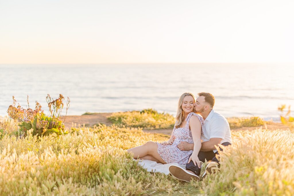 Groom kissing bride sitting in a grass field on a cliff in Carlsbad.