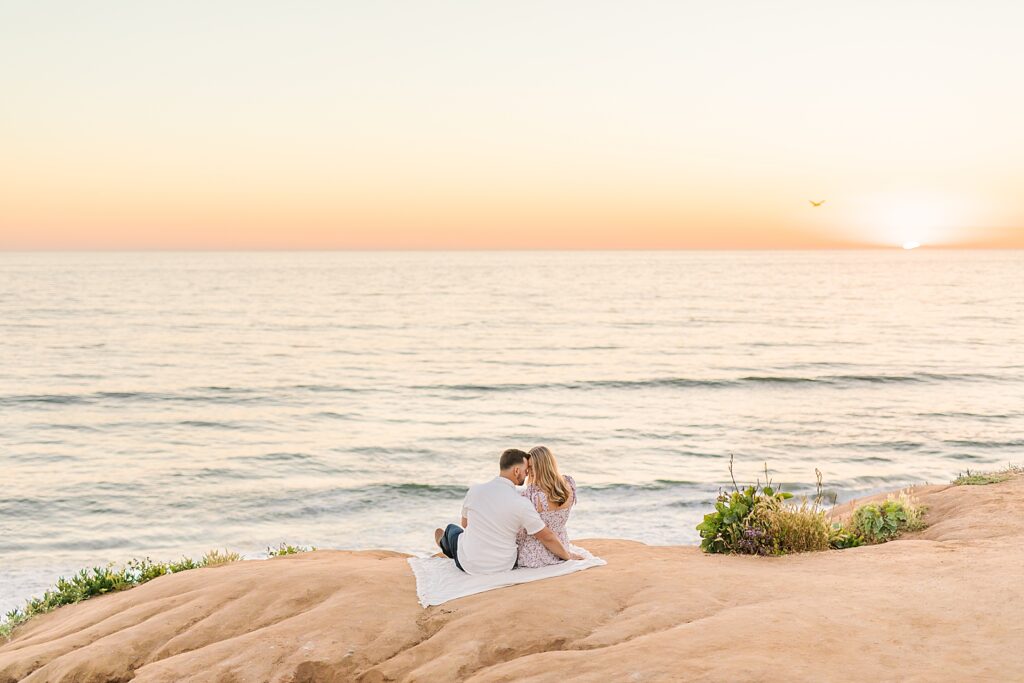 Couple cuddling on cliff in Carlsbad as sun sets behind ocean by Sherr Weddings.