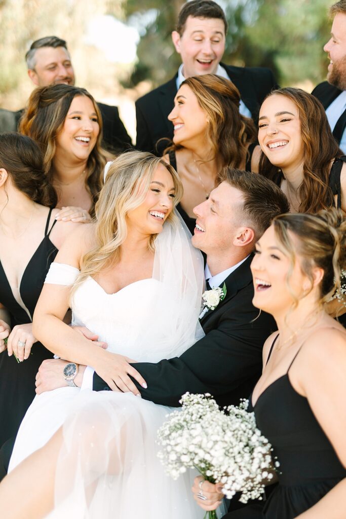 Bride and groom sitting together among their bridal party at Lake Oak Meadows in Temecula, California.
