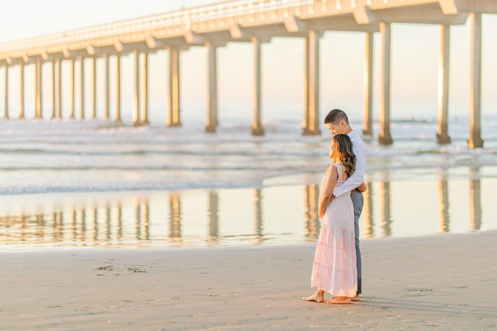 Asian couple enjoying a maternity photo session at Scripps Pier in La Jolla, with the woman wearing a pink ruffled dress and the man in a white button-down shirt and pants, standing by the beach at sunset.