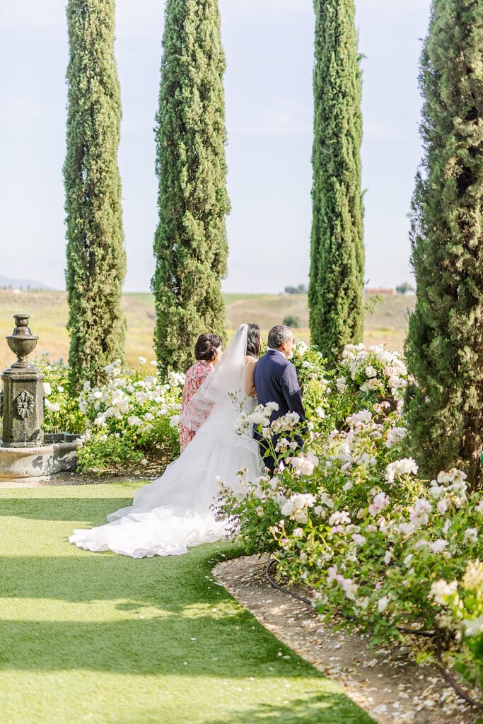 Candid photo of bride walking arm in arm with her parents as she makes her way down the ceremony aisle, the lush flowers and trickling fountain around her, at Falkner Winery in Temecula, California.