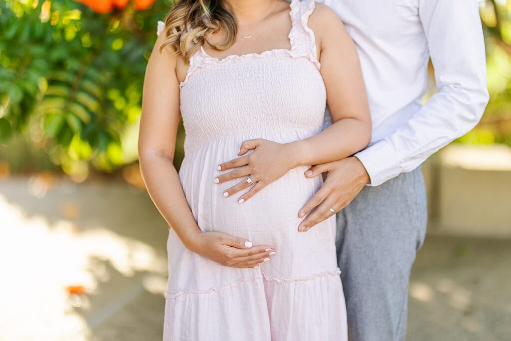 Asian couple captures a magical maternity moment at Scripps Pier in La Jolla. The woman is dressed in a pink ruffled gown, and the man is in a white shirt and pants, standing near the water with the pier in the background.