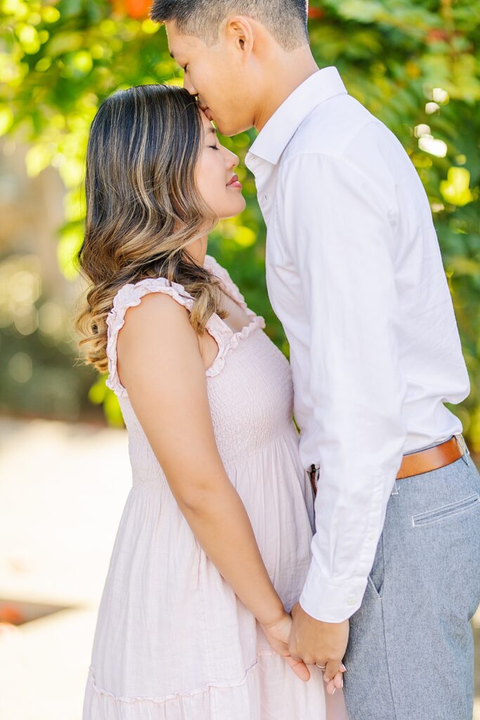 Asian couple enjoying a maternity photo session at Scripps Pier in La Jolla, with the woman wearing a pink ruffled dress and the man in a white button-down shirt and pants, standing by the beach at sunset.