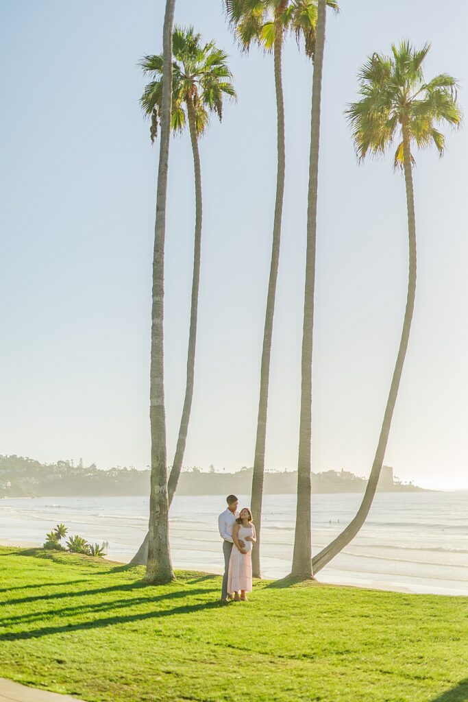 A couple in a pink ruffled maternity dress and a white button-down shirt pose together by Scripps Pier in La Jolla, with the beach and soft golden hour lighting in the background.