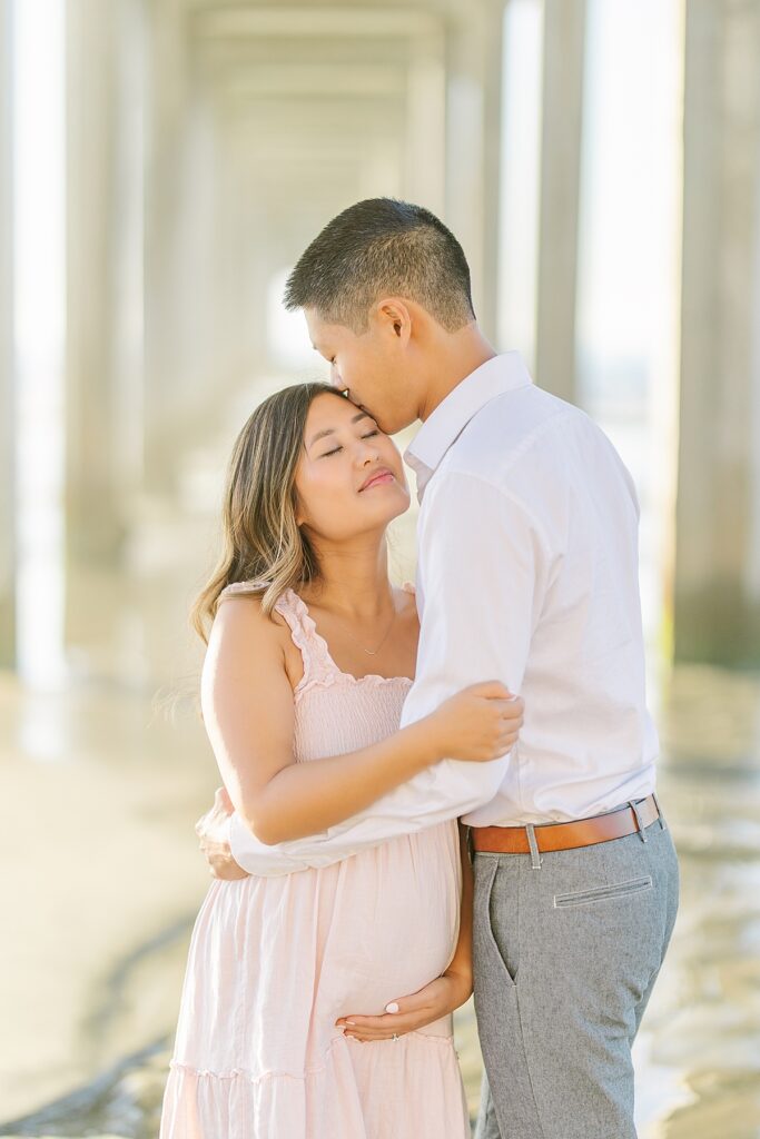 Asian couple captures a magical maternity moment at Scripps Pier in La Jolla. The woman is dressed in a pink ruffled gown, and the man is in a white shirt and pants, standing near the water with the pier in the background.