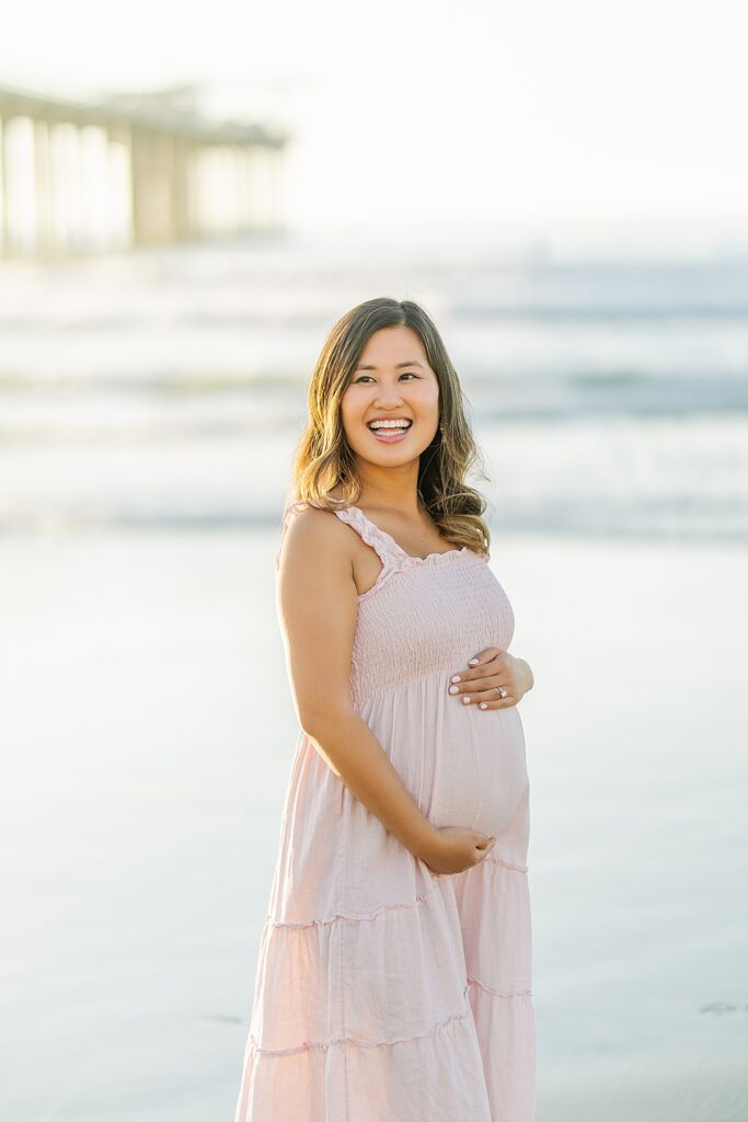 A couple in a pink ruffled maternity dress and a white button-down shirt pose together by Scripps Pier in La Jolla, with the beach and soft golden hour lighting in the background.