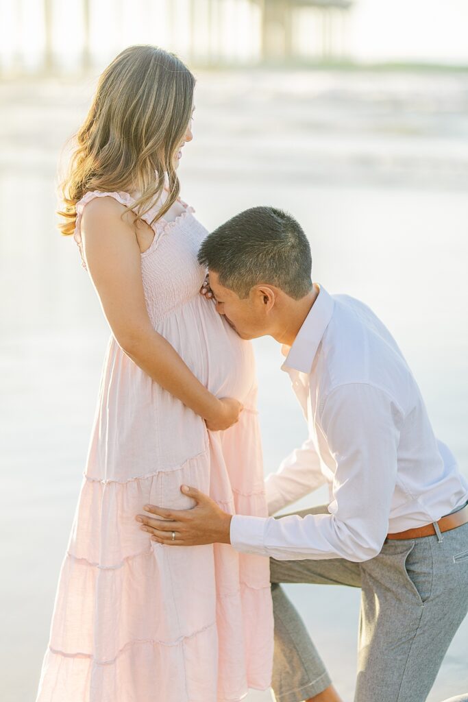 Asian couple enjoying a maternity photo session at Scripps Pier in La Jolla, with the woman wearing a pink ruffled dress and the man in a white button-down shirt and pants, standing by the beach at sunset.