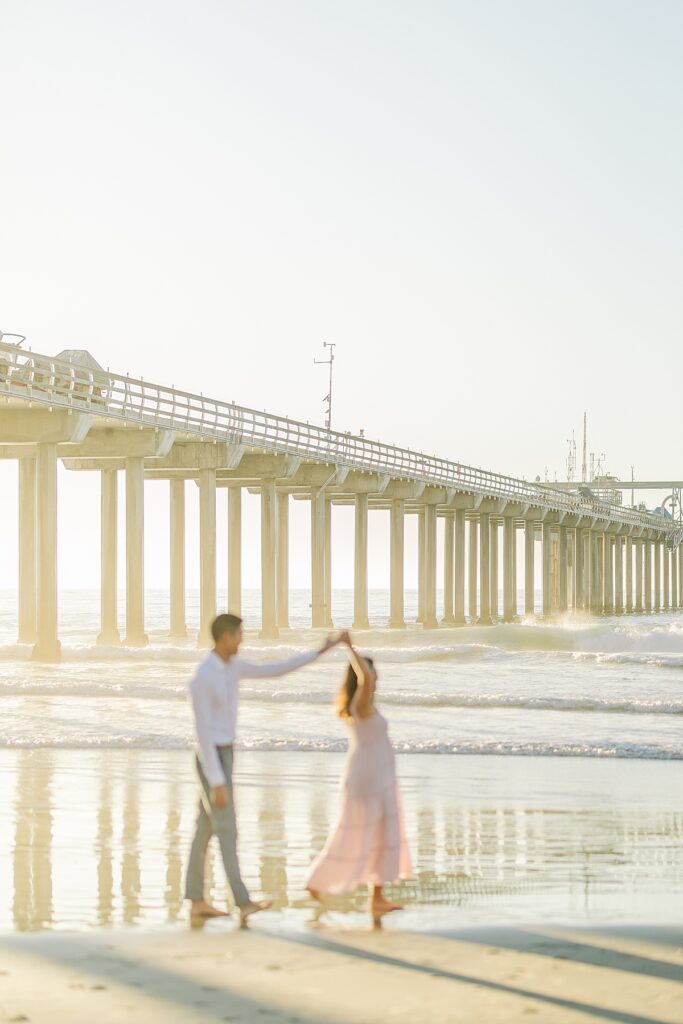 Asian couple captures a magical maternity moment at Scripps Pier in La Jolla. The woman is dressed in a pink ruffled gown, and the man is in a white shirt and pants, standing near the water with the pier in the background.