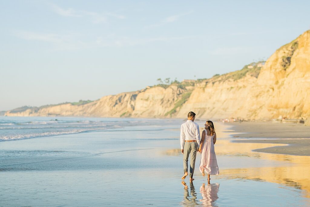 A couple in a pink ruffled maternity dress and a white button-down shirt pose together by Scripps Pier in La Jolla, with the beach and soft golden hour lighting in the background.