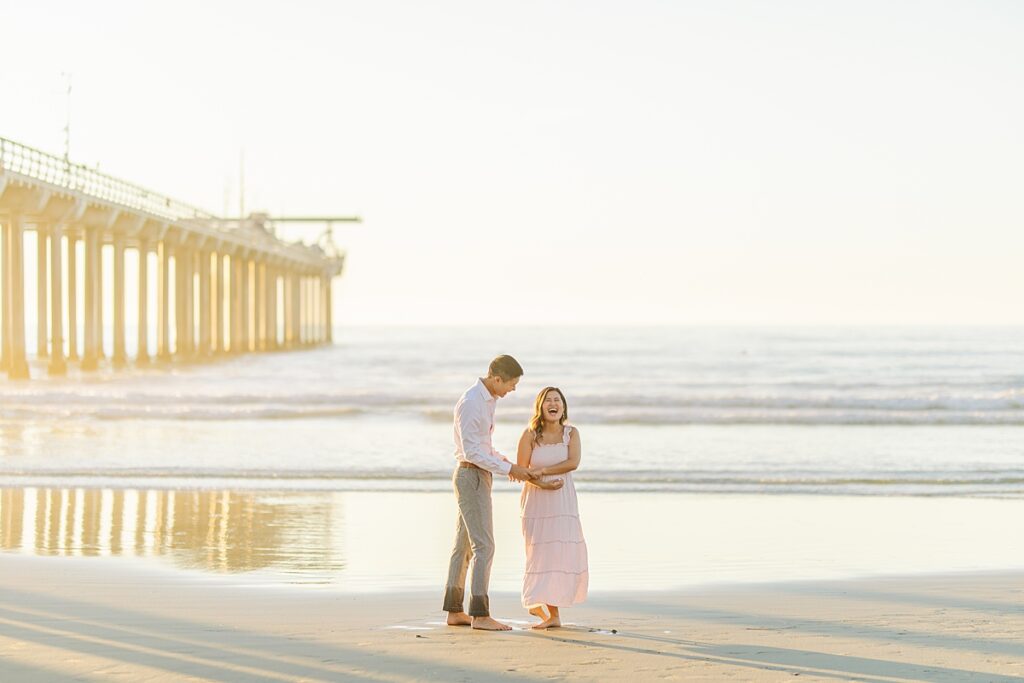 A couple in a pink ruffled maternity dress and a white button-down shirt pose together by Scripps Pier in La Jolla, with the beach and soft golden hour lighting in the background.