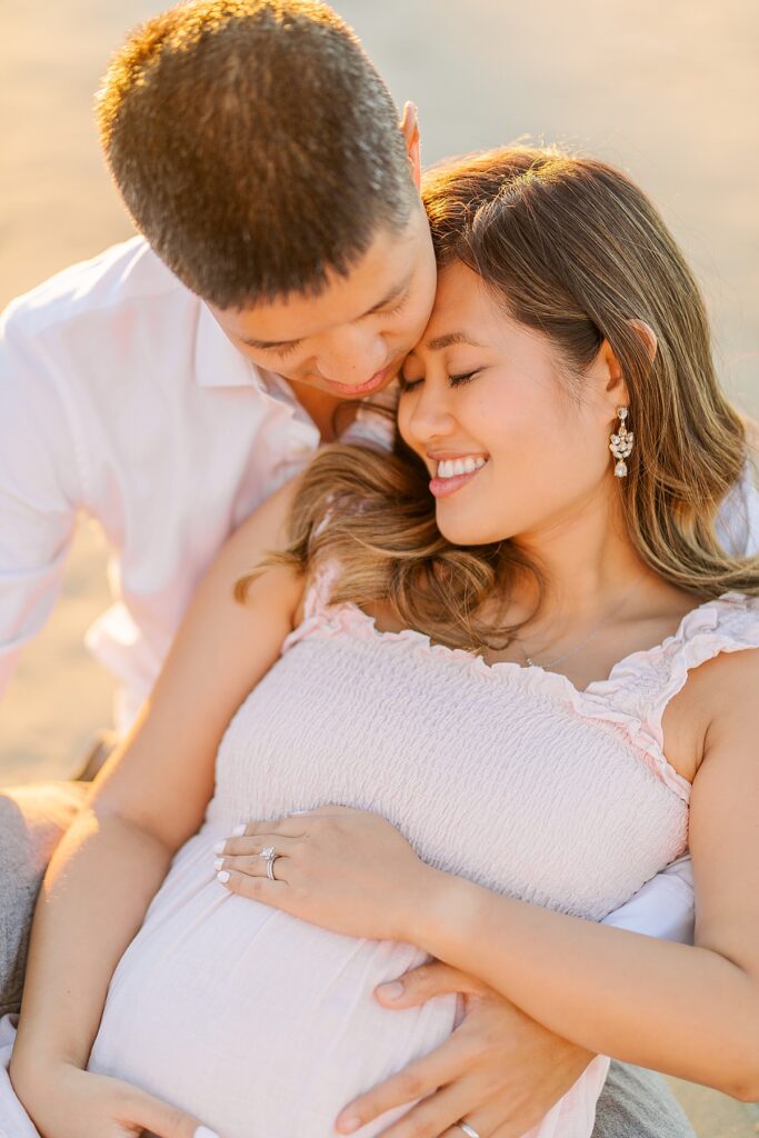 Maternity photos of an Asian couple at Scripps Pier in La Jolla. The expectant mother wears a pink ruffled dress, while the father-to-be is dressed in a white button-down shirt and pants. They’re standing on the beach, surrounded by soft, golden light.
