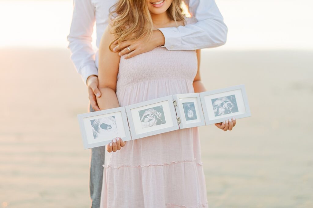Maternity photos of an Asian couple at Scripps Pier in La Jolla. The expectant mother wears a pink ruffled dress, while the father-to-be is dressed in a white button-down shirt and pants. They’re standing on the beach, surrounded by soft, golden light.