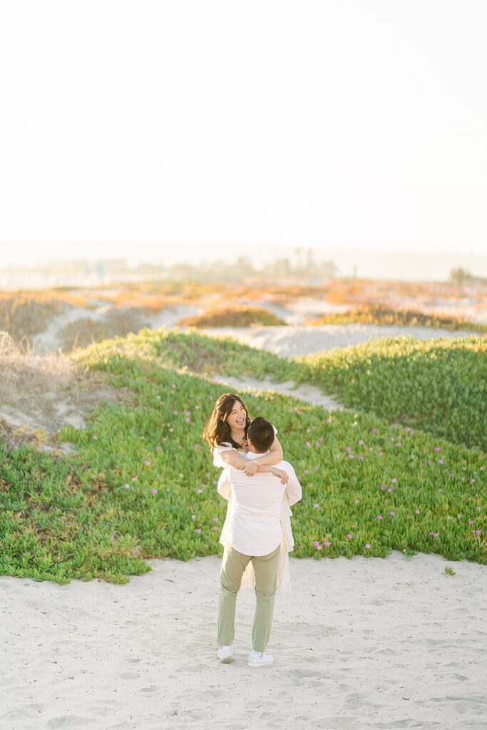 Jiaqi and Eric share a joyful laugh in the sand dunes, playfully embracing each other as they enjoy their engagement session in golden light.