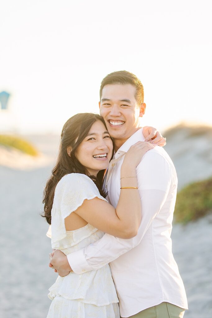 Couple laughing together and smiling at the camera during their beach engagement session at Coronado Beach in San Diego, California.