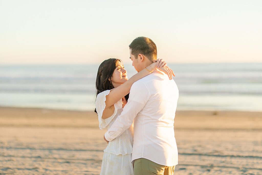 Jiaqi and Eric stand together on the beach, bathed in soft golden light, gazing lovingly at each other as the waves gently crash in the background.