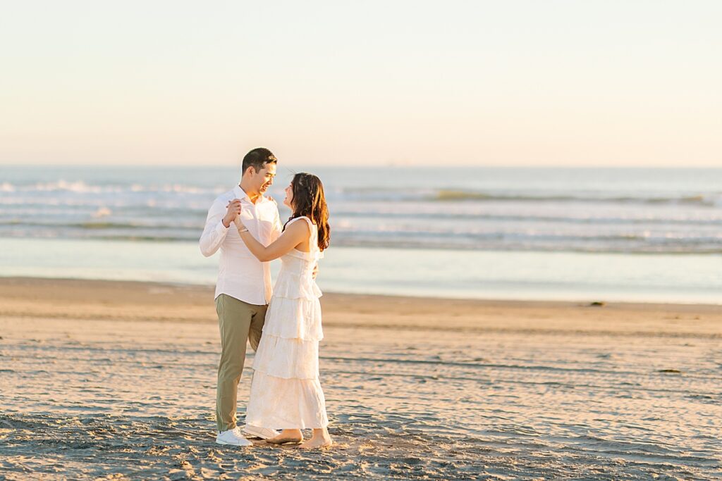 Jiaqi and Eric stand together on the beach, bathed in soft golden light, gazing lovingly at each other as the waves gently crash in the background.