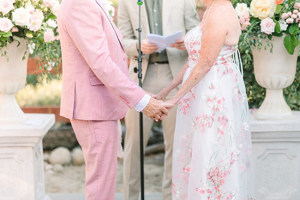 Bride in white dress with pink and green florals and groom in blush pink suit holding hands during their wedding ceremony in Laguna Hills, California.