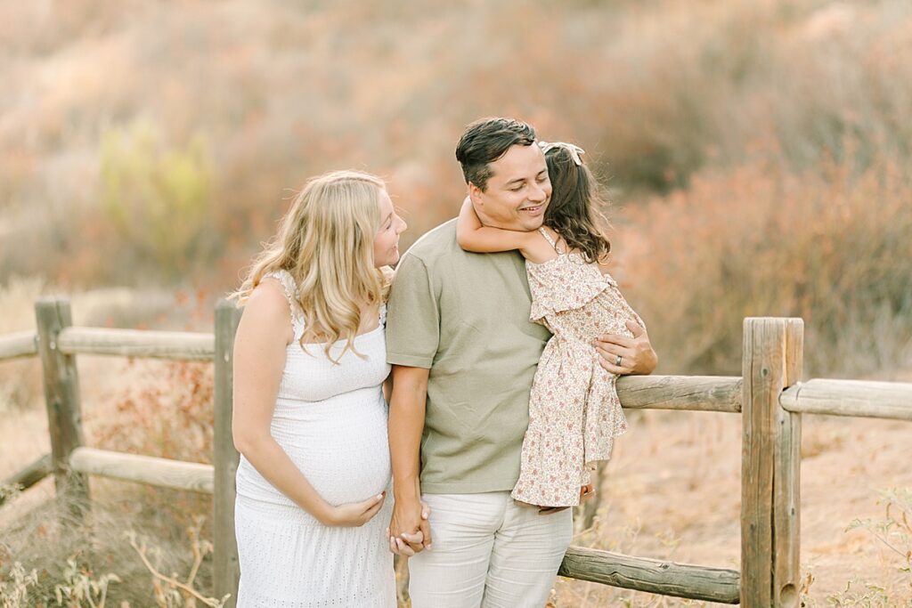 Toddler daughter hugging her dad while he holds moms hand on a wooden fence in the wilderness.