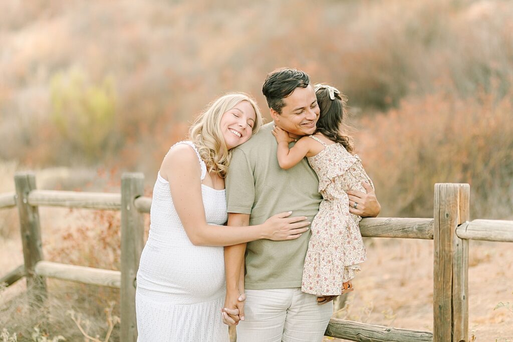 Family of three sharing a big hug before welcoming their new baby on a fence, smiling together.