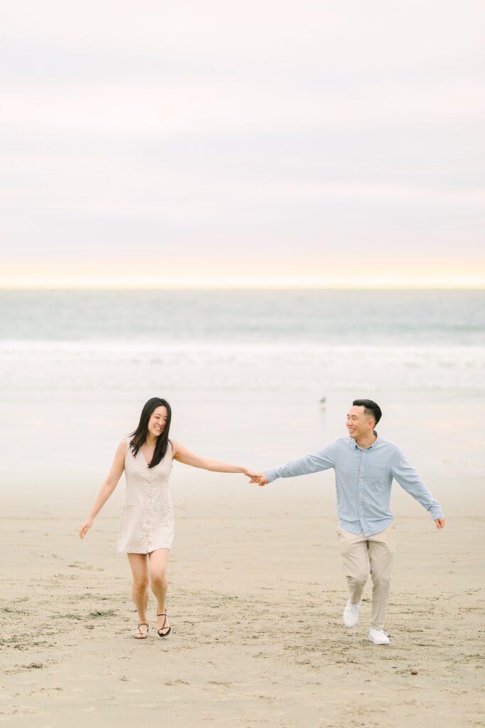 Engaged couple walking along Carlsbad Beach during a pink and purple sunset.