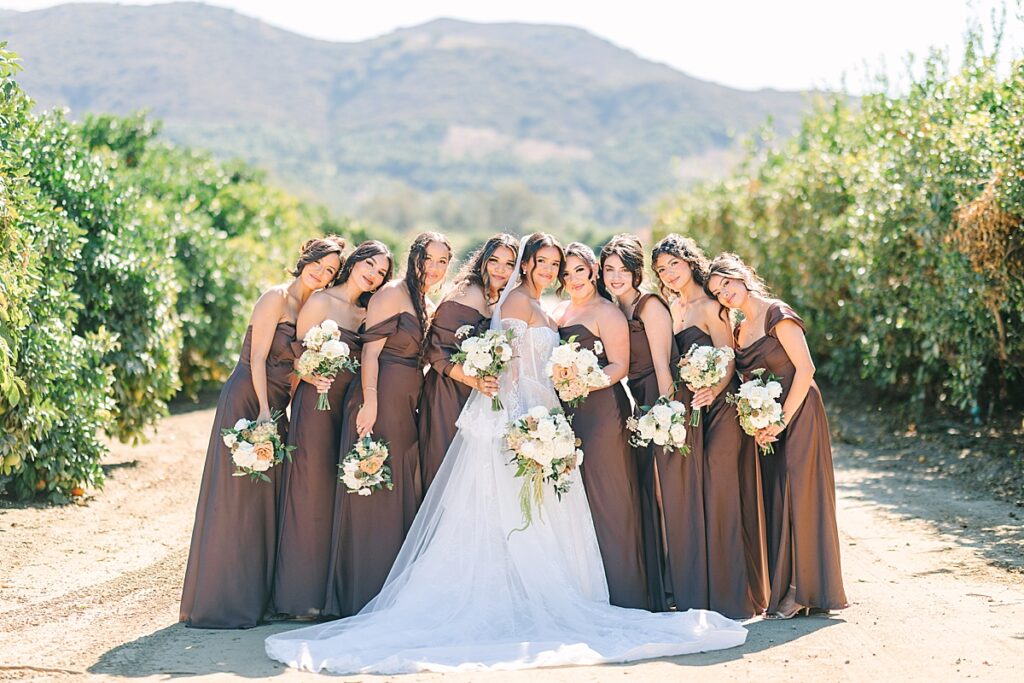 Bride and her bridesmaids leaning in for a hug in the middle of orange orchards at Rancho Guejito Vineyard in Escondido.