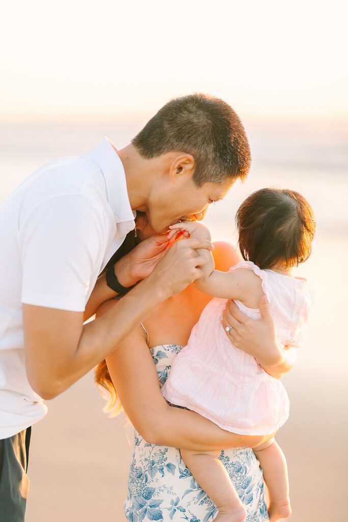 Dad giving his baby girl a kiss on the hand while mom holds her at their beach family photography session at Del Mar Beach.